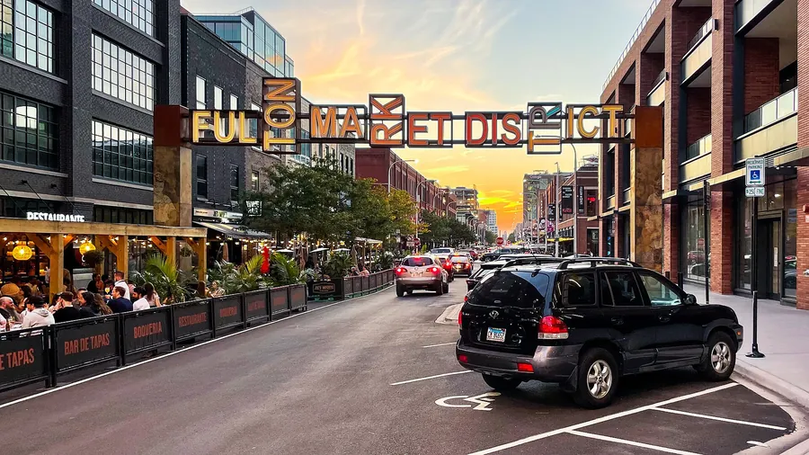 Fulton Market in 2021 at sunset: the Fulton Market District sign arching over the street, with crowded outdoor dining terraces and converted warehouse buildings on either side.