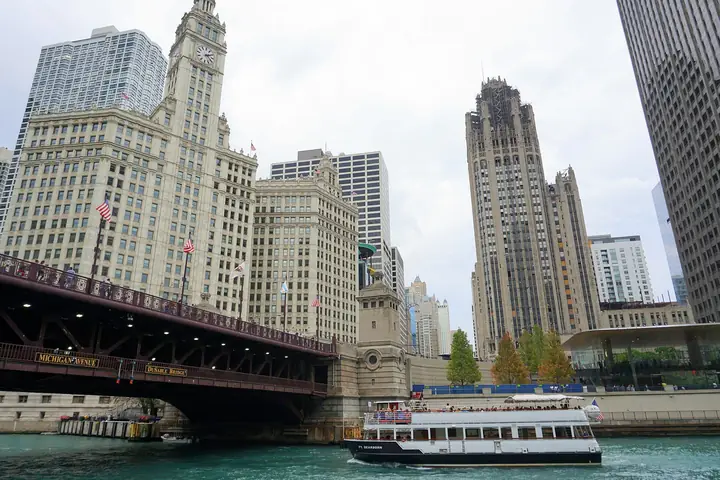 The white terracotta Wrigley Building and the Tribune Tower rising above the DuSable Bridge and the Chicago River, with a tour boat below.