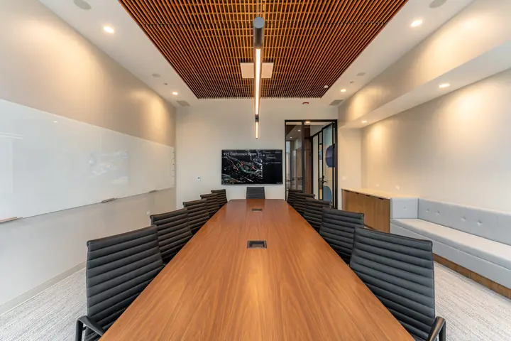 A boardroom with a long walnut table, black leather chairs, a slatted wood ceiling and a wall-mounted display.