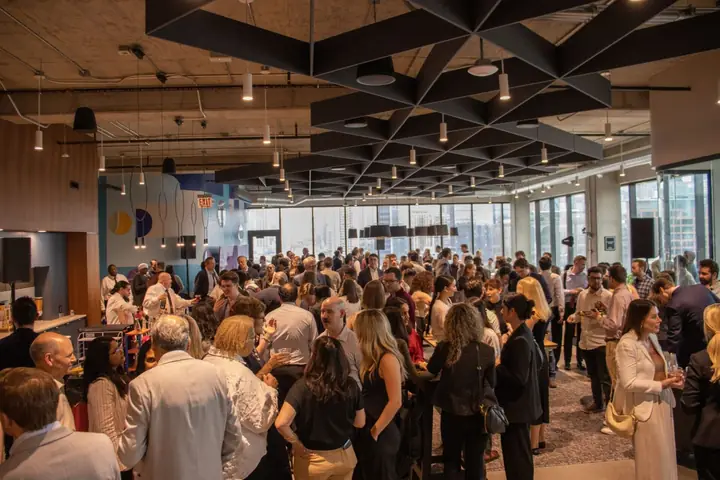 A packed evening reception under a geometric black ceiling in Portal's common area, with the city skyline in the windows behind.