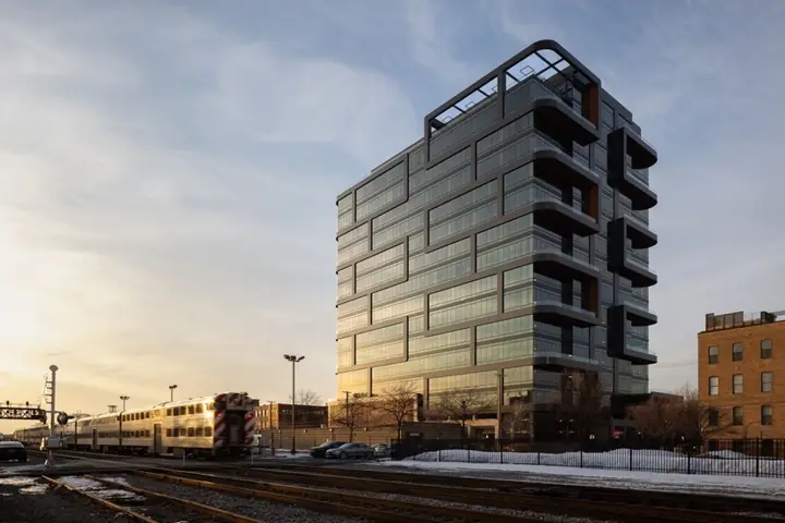 Fulton Labs at sunset: a glass and dark-metal tower with stacked, cantilevered balconies, beside the Metra tracks with a commuter train passing.