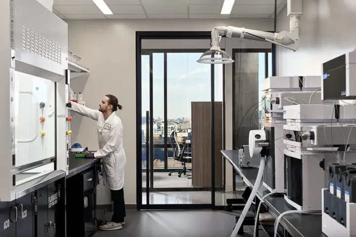 A scientist working at a chemical fume hood in a Portal lab, with analytical instruments to the right and floor-to-ceiling windows overlooking the city.