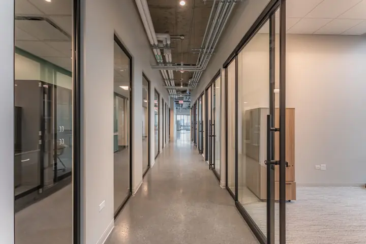A long, bright corridor of black-framed glass doors and walls leading to lab and office suites.