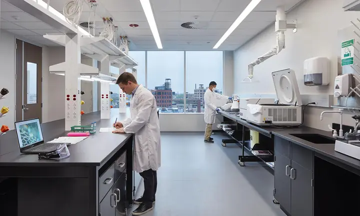 A scientist in a lab coat writes at a black lab bench in a bright Portal lab, with a second scientist at the instruments behind him and a wall of windows looking out over the brick rooftops of the West Loop.