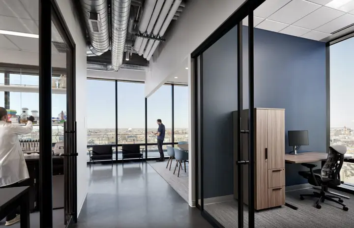 Glass-walled private offices with sit-stand desks and wide windows over the city.