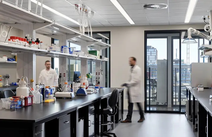 Two scientists in lab coats at long lab benches stocked with reagents and equipment, beside tall windows.