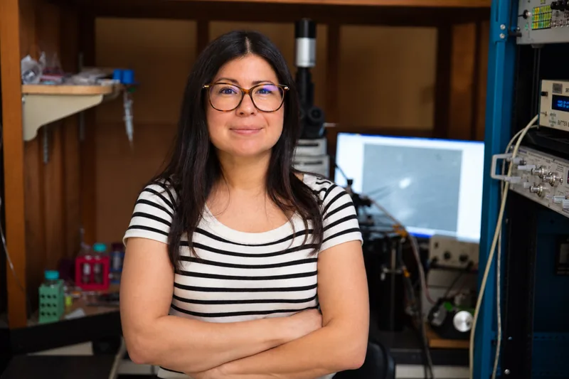 Dr. Melissa Hernandez, in glasses and a striped top, arms folded, in her lab at the Salk Institute, with a microscope and electrophysiology rig behind her.