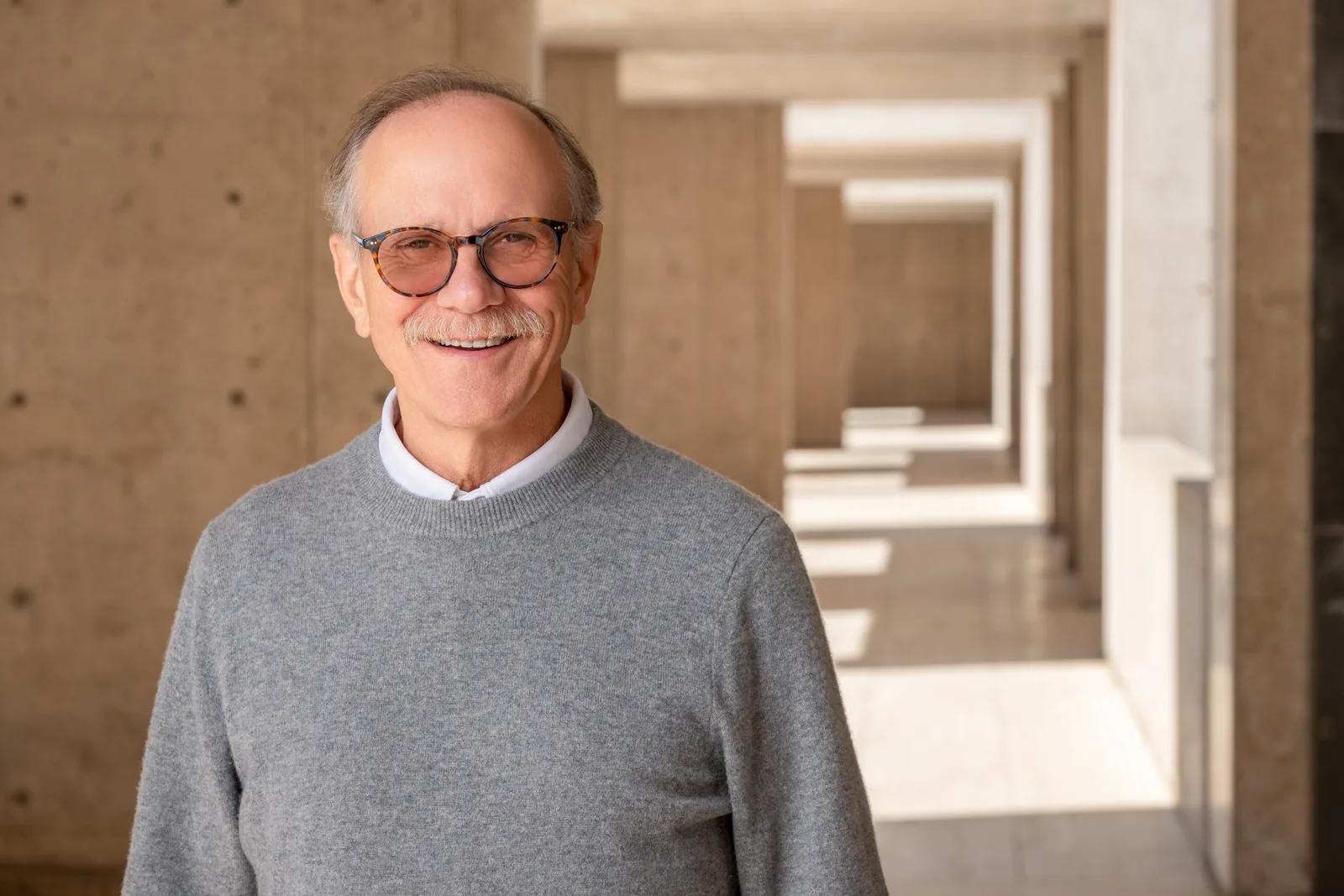 Dr. Rusty Gage, smiling, in a gray sweater and glasses, in a sunlit concrete corridor at the Salk Institute.