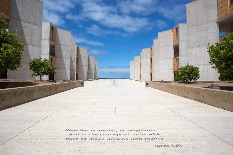 The Salk Institute courtyard under a blue sky, its travertine plaza running between the concrete laboratory towers toward the Pacific. Jonas Salk's words are carved into the stone: Hope lies in dreams, in imagination and in the courage of those who dare to make dreams into reality.