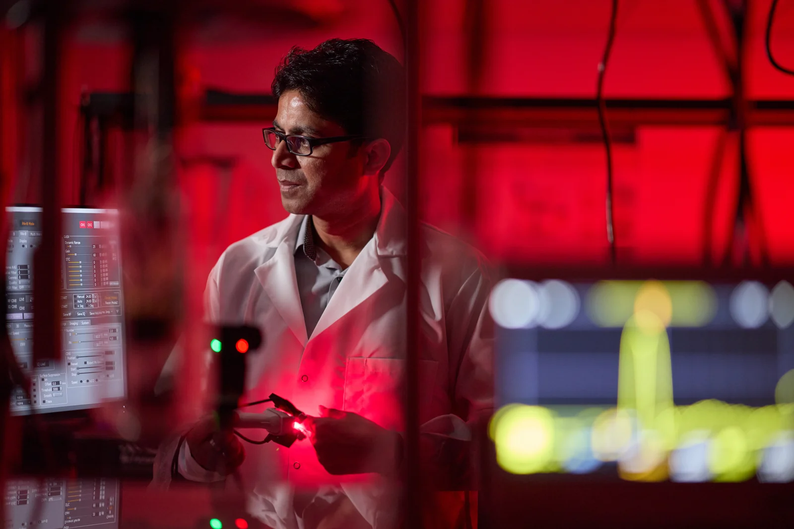Dr. Manojit Pramanik at work in the Biomedical Imaging Laboratory, lit by the red glow of the laser lab, holding a fiber-coupled probe beside an instrument rack and acquisition monitors.